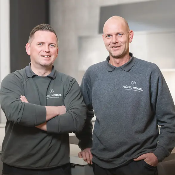 Zwei Männer stehen nebeneinander im Möbelhaus, tragen Pullover mit Aufdruck Möbel Hensel und verschränken die Arme